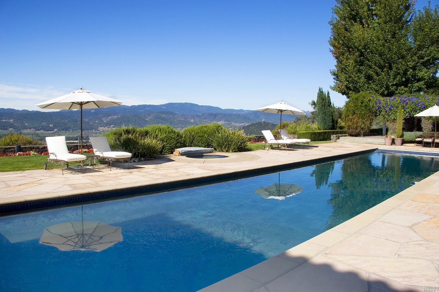 A long lap pool flanked by stone pavers and white umbrellas, looking west toward the Napa valley floor and the Mayacamas Mountains beyond.