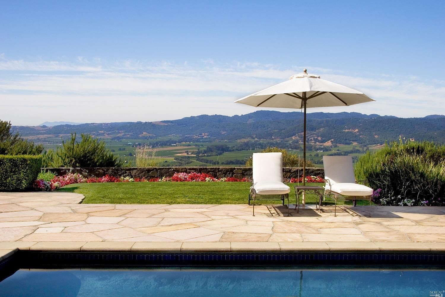 Two chaise lounges and a white umbrella beside the pool, a flagstone terrace in the foreground and the valley falling away behind.