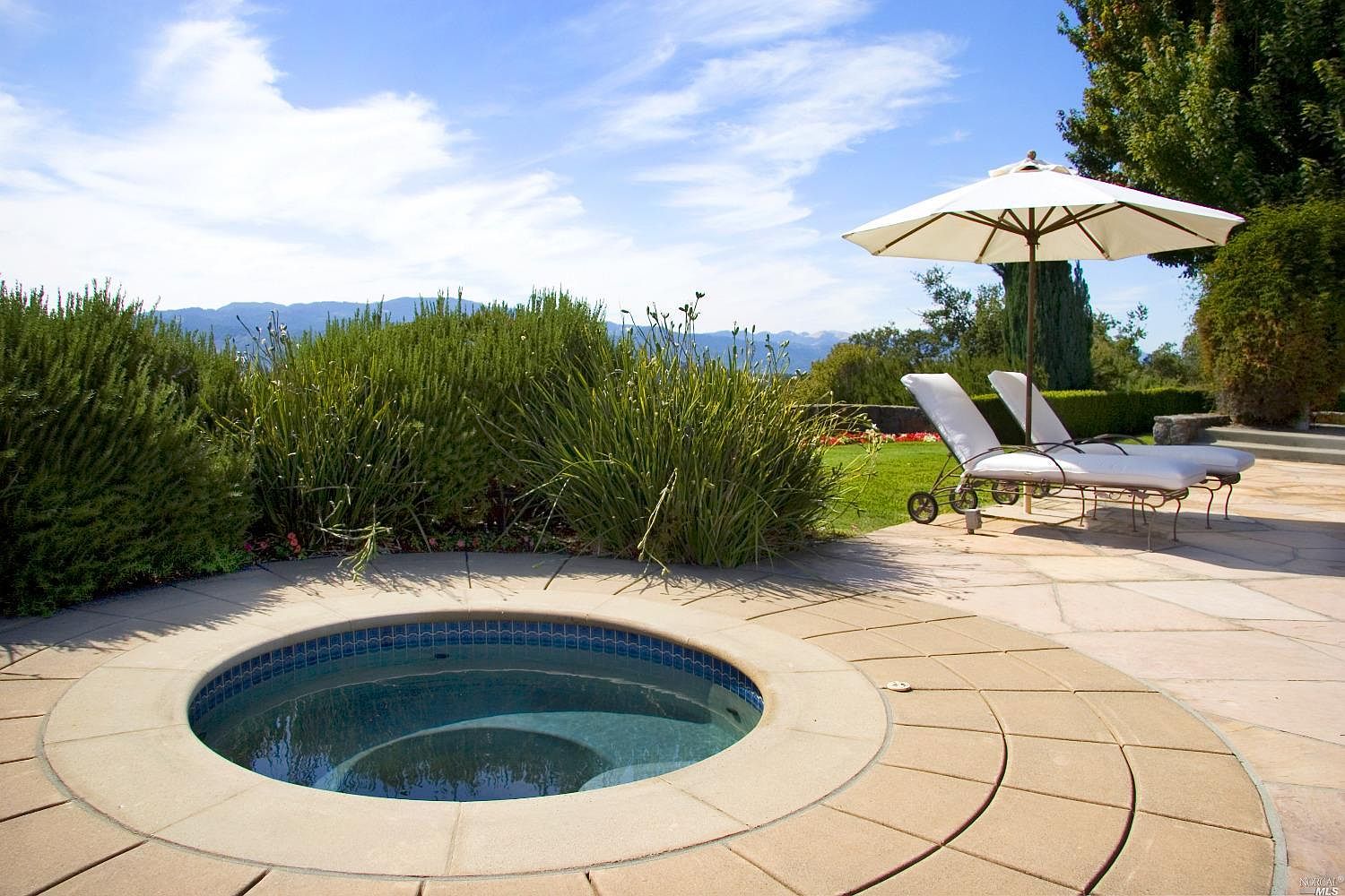 A round spa set into a flagstone patio next to garden plantings, with a chaise lounge under a white umbrella beyond.